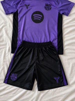 Lila och svart FC Barcelona träningsset - Snyggt träningsset med lila t-shirt och svarta shorts. T-shirten har FC Barcelona-logga, Kobe Bryant-symbol och Spotify-tryck framtill. Shortsen är svarta med lila detaljer och har samma loggor. Perfekt för fotboll eller gymmet.