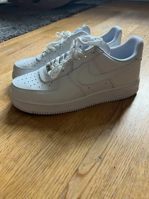 Vita Nike Air Force 1 sneakers - Klassiska helvita Nike Air Force 1 sneakers i läder med perforerad tå, vita skosnören och den ikoniska swoosh-loggan på sidan. Tjock sula och låg profil gör dem perfekta för en clean och stilren look.