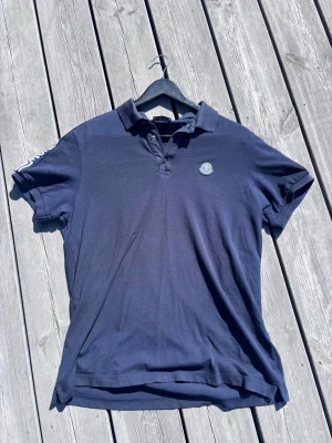 Marinblå pikétröja från Moncler - Snygg marinblå pikétröja från Moncler med klassisk krage och knappar framtill. Tröjan har korta ärmar och en diskret Moncler-logga på bröstet samt randiga detaljer med Moncler-tryck på axeln. Perfekt till en avslappnad men stilren look.