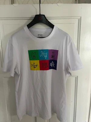 Vit t-shirt - Vit t-shirt från Ginger med ett stort färgglatt tryck på bröstet i grönt, blått, lila, gult, rött och mörkblått. Trycket består av sex rutor med abstrakta illustrationer och text. Klassisk passform och rund hals.