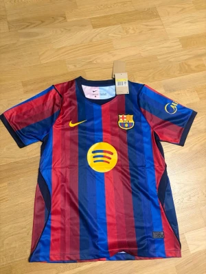 Barca 26/27 hemma - Säljer en officiell FC Barcelona matchtröja från Nike med klassiska blå och vinröda ränder. Tröjan har klubbmärke, Nike-logga och Spotify-logga på bröstet samt korta ärmar. Tillverkad i lätt och ventilerande polyester, perfekt för fotbollsträning eller match.
