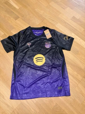 Lamine yamal borta 26/27 S - Snygg Barcelona matchtröja i svart och lila med gula detaljer. Tröjan har korta ärmar, klubbmärke på bröstet, Spotify-logga och Lamine Yamal samt nummer 10 på ryggen. Perfekt för dig som älskar fotboll och vill stötta Barca.