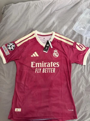 Real Madrid 26/27 Mbappe version - Säljer en vinröd Real Madrid bortatröja från Adidas med vita detaljer och tryck. Tröjan har Mbappé och nummer 10 på ryggen, samt klubbmärke och sponsorlogga framtill. Korta ärmar med vita ränder på axlarna. Perfekt för fotbollsfans!