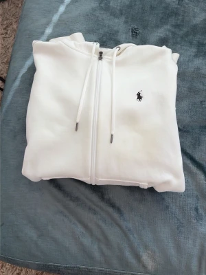 Vit hoodie med dragkedja från Polo Ralph Lauren - Säljer en stilren vit hoodie från Polo Ralph Lauren med dragkedja framtill, huva med dragsko och två fickor. Klassisk logga broderad på bröstet. Tillverkad i mjukt material som känns skönt mot huden. Perfekt för en avslappnad och trendig look.