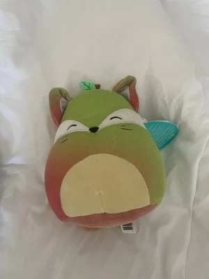 Grön och brun Squishmallow räv - Mysig Squishmallow i form av en räv med grön och brun färg, vita detaljer vid ögonen och en liten grön svans med brun spets. Mjuk plysch och rund form, perfekt som gosedjur eller prydnad. Har små öron och ett broderat ansikte.