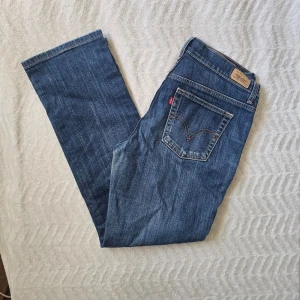 Blå Levi's raka jeansW28L30 - Klassiska blå jeans från Levi's med rak passform och fem fickor. Jeansen har den ikoniska röda Levi's-lappen på bakfickan och patch med logga i midjan. Tillverkade i slitstark denim med kontrastsömmar. Perfekta för en avslappnad och tidlös stil.