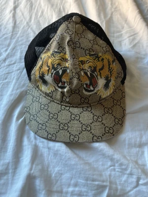 Beige keps med tigertryck och nät - Snygg beige keps med GG-mönster och två rytande tigrar framtill. Kepsen har svart nät på sidorna och justerbar rem bak. Perfekt för dig som vill sticka ut med en unik accessoar.