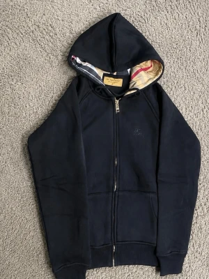 Svart  hoodie med dragkedja Burberry - Snygg svart hoodie från Burberry med dragkedja framtill och klassiskt rutigt foder i huvan. Hoodien har fickor på magen, ribbade muddar och diskret broderad logga på bröstet. Perfekt för en chill och stilren look. Den är Size S men jag skulle säga att den passar Xs eftersom burberry hoodies är gjorda för att sitta tight och skönt så jag skulle rekommendera den för dig om due mellan 155-170