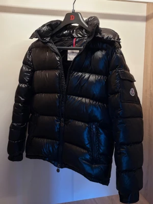 Moncler Maya Dunjacka (Svart) - Snygg svart dunjacka från Moncler med glansig finish, huva och dragkedja framtill. Dunjackan har två pytte små hål på den högre armen längst upp, knappt synliga om man inte kollar jätte nära. Annars är allt perfekt. Jackan är i storlek S, passar lite som M också. Jackan är äkta, kvitto finns.
