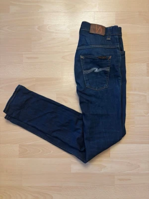 Nudie Jeans - Marinblå  - Snygga mörkblå Nudie Jeans i klassisk modell. Stilren mörk denim med orange sömmar och den klassiska Nudie-detaljen på bakfickan. Mycket fint skick. 📏 Storlek: W30 L34 👖 Färg: Mörkblå 📦 Skickas snabbt