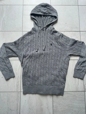 Grå hoodie Polo Ralph Lauren - Snygg grå kabelstickad hoodie från Polo Ralph Lauren med dragsko i huvan och broderad logga på bröstet. Tröjan har långa ärmar, ribbade muddar och är gjord i mjukt material som passar perfekt till höst och vinter.