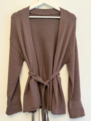 Stickad cardigan - Mjuk brun cardigan utan knäppning framtill. Koftan har ett tillhörande knytband i midjan för en smickrande silhuett, nedhasade axelsömmar och breda ribbstickade manschetter vid ärmsluten. Perfekt plagg för lager-på-lager.