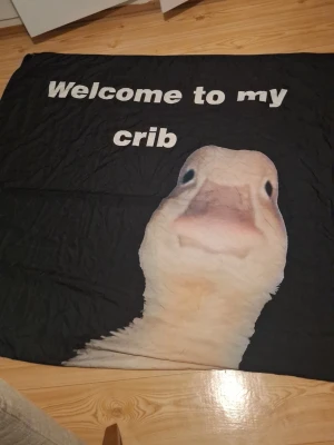 Svart Duk med anka och text - Rolig svart duk med stor bild på en anka och texten 'Welcome to my crib' i vitt. Perfekt att sätta upp på väggen , de tillkommer två krokar. Mjuk och lätt material, passar dig som gillar humoristiska detaljer i hemmet.