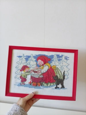 Broderad tavla med vintermotiv - Broderad tavla i röd ram med vintermotiv som föreställer två barn och en katt i snölandskap. Tavlan har broderade detaljer i rött, grönt, blått och vitt. Perfekt som dekoration för dig som gillar retro och handarbete.