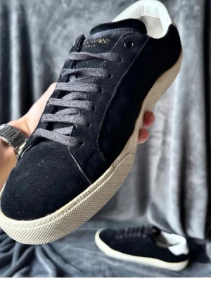 Saint Laurent skor - Stilrena svarta sneakers i mocka med vit sula och vit detalj på hälen. Skorna har svarta skosnören och en klassisk låg profil. Perfekta för dig som gillar en clean och tidlös look.