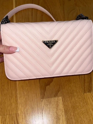 Ljusrosa  handväska crossbody - Supersnygg ljusrosa handväska med quiltat mönster och silverfärgade detaljer. Väskan har en stilren rektangulär form och ett klassiskt metallogo framtill. Perfekt accessoar för att lyfta din outfit.