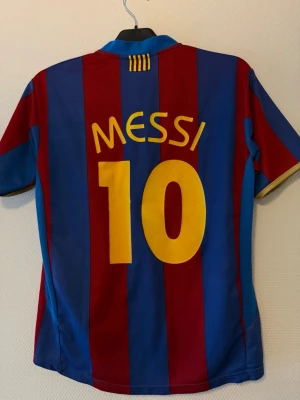Barcelona Messi matchtröja - Säljer en klassisk FC Barcelona matchtröja med Messi och nummer 10 på ryggen. Tröjan har blå och vinröda vertikala ränder, gul text och klubbmärke på bröstet. Tillverkad i lätt och ventilerande material, perfekt för fotboll eller som supporterplagg.