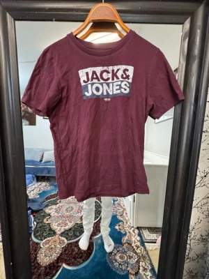 Vinröd Jack & Jones t-shirt - Säljer en vinröd t-shirt från Jack & Jones med tryckt logga i vitt och grått på bröstet. T-shirten har rund halsringning och korta ärmar. Perfekt till en avslappnad stil och enkel att matcha med jeans eller shorts.