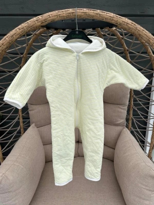 Barnoverall strl. 74 - Mysig overall i ljusgul och vit randig design med dragkedja framtill och huva. Perfekt för att chilla hemma eller som sovplagg. Tillverkad i mjukt tyg.