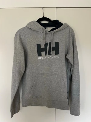 Grå Helly Hansen hoodie med huva - Säljer en grå hoodie från Helly Hansen med stor svart HH-logga och text på bröstet. Hoodien har huva med dragsko och en stor magficka. Tillverkad i mjukt sweatshirtmaterial, perfekt för chill dagar.