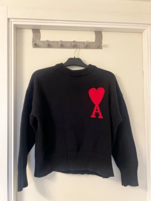 Ami paris stickat tröja - Svart stickad tröja med rund hals och ribbade muddar. På bröstet finns ett stort rött hjärta och bokstaven 'A' i samma färg. Tröjan har en avslappnad passform och är perfekt för dig som gillar stilrena detaljer.