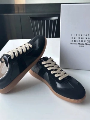 Svarta sneakers Maison Martin Margiela - Snygga svarta sneakers från Maison Martin Margiela med vita snören och gummisula i beige. Skorna har en klassisk låg siluett och är tillverkade i en mix av läder och mocka för en stilren look.