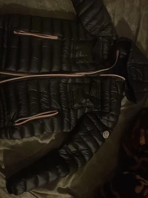 Svart pufferjacka med huva - Snygg svart pufferjacka med huva och quiltad design. Jackan har dragkedja framtill och två sidofickor med kontrastfärgade detaljer. Perfekt för kalla dagar och ger en sportig look.