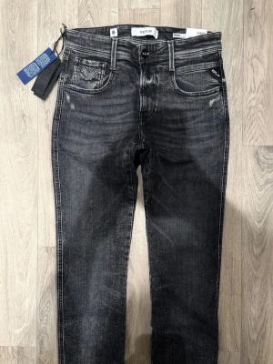 Helt Nya Replay Anbass Aged 05 Year Mörkgrå W30/L32 - Slim Fit - Replay Anbass Aged 05 Year mörkgrå jeans i organic cotton med snygga slitningar och slim fit-passform som sitter smalt och bekvämt, storlek W30/L32, midja 38 cm, ytterbenslängd 109 cm, benöppning 15,5 cm, modellen är 185cm & väger 65kg, Helt Nya Med Alla Tags.