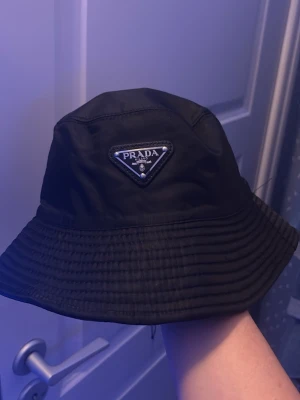 Svart bucket hat från Prada - Snygg svart bucket hat från Prada med den ikoniska trekantiga logotypen framtill. Hatten är tillverkad i ett glansigt, slitstarkt material som ger en clean och modern look. Perfekt accessoar för att lyfta din outfit och ge en exklusiv touch.