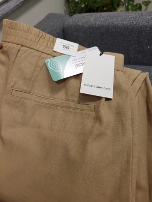 Beige chinos från These Glory Days - Snygga beige chinos från These Glory Days med elastisk midja och raka ben. Byxorna har diskreta sidofickor och en bakficka, samt en extra ficka på benet. Perfekta för en avslappnad men stilren look.