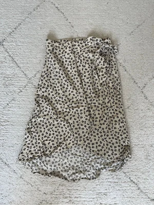 Beige leopardmönstrad omlottkjol - Säljer en beige omlottkjol med svart leopardmönster och knyt i midjan. Kjolen har en lätt och luftig känsla, perfekt för varmare dagar. Den har en något asymmetrisk nederkant och är enkel att styla med både sneakers och sandaler.
