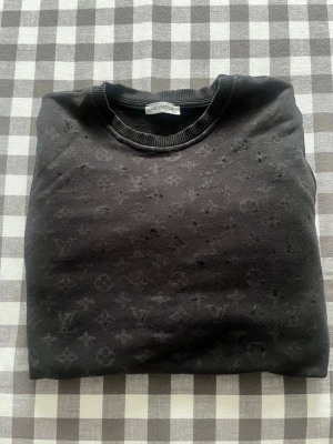Svart Louis Vuitton sweatshirt - Snygg svart sweatshirt från Louis Vuitton med diskret monogrammönster över hela tröjan. Rund halsringning och ribbade muddar ger en klassisk look. Perfekt för dig som gillar exklusiva streetwear-plagg och vill sticka ut med stil.