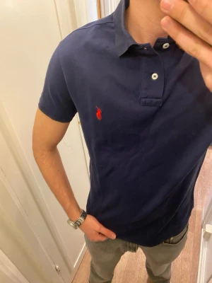 Mörkblå Piké från Polo Ralph Lauren - Klassisk marinblå pikétröja från Ralph Lauren med korta ärmar, krage och två knappar framtill. Tröjan har den ikoniska röda broderade loggan på bröstet och är gjord i mjuk bomull som känns skön mot huden. Enkel att matcha till olika stilar. Storlek Large. Snabb Leverans!