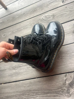 Svarta lackade Dr Martens - Säljer ett par svarta kängor i glansigt lackmaterial med klassisk snörning och gul söm runt sulan. Kängorna har en robust sula och dragflik bak. Perfekta för dig som vill ha en edgy och stilren look.