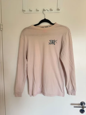 Ljusrosa långärmad topp med tryck - Fnatic - Ljusrosa långärmad topp med en liten svart grafisk logga på bröstet och en stor grön tecknad fladdermus på ryggen. Rund halsringning och mjukt material som känns skönt mot huden. Perfekt för dig som gillar streetwear och coola prints. Fnatic.