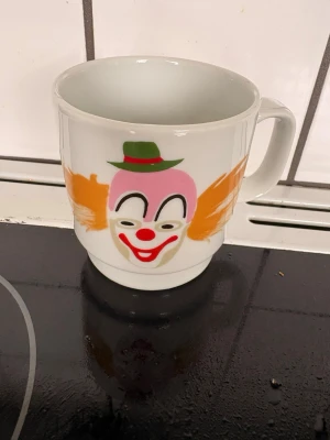 Mugg med clownmotiv från B&G - Unik vit porslinsmugg från B&G med färgglatt clownansikte på framsidan. Clownen har rosa ansikte, röd näsa, grönt hatt och orange hår. Perfekt för dig som gillar retro eller vill ha något annorlunda i köksskåpet.