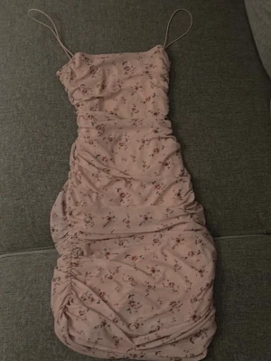 Ljusrosa blommig klänning NLY One - Söt ljusrosa kortklänning från NLY One med smala axelband och blommigt mönster. Klänningen har rynkade detaljer längs sidorna och är figurnära med en feminin siluett. Perfekt för sommarens alla tillfällen.