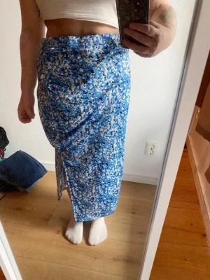 Blommig blå omlottkjol med slits - Säljer en lång omlottkjol i blått med vitt och svart blommönster. Kjolen har en snygg slits på sidan och knyts i midjan för en skön och justerbar passform. Perfekt för dig som gillar färg och mönster!