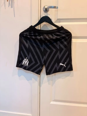 Svarta Marseille shorts från Puma - Snygga svarta träningsshorts från Puma med Marseille-logga och texten 'Droit au but' på vänster ben. Shortsen har ett grått blixtmönster och elastisk midja för en skön passform 