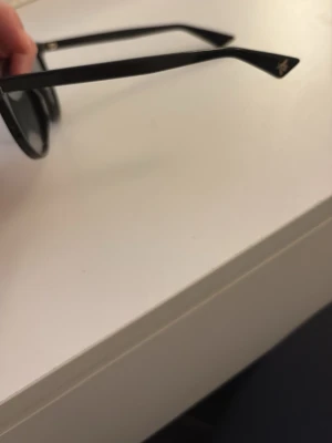 Svarta runda solglasögon - Snygga svarta solglasögon med rundad form och mörka glas. Bågarna är tunna och har små guldfärgade detaljer vid gångjärnen. Perfekta för att höja din stil på soliga dagar.