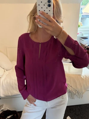 Lila blus med veckad framsida - Snygg lila blus med rund halsringning och en liten öppning framtill. Blusen har veckade detaljer på framsidan och långa ärmar med knappar vid ärmsluten. 