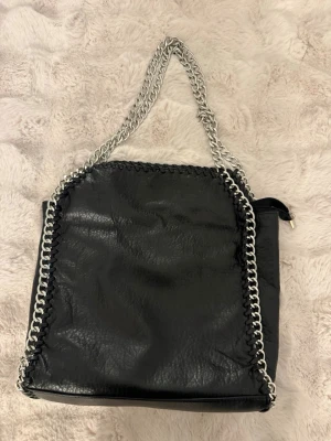Tiamo väska i L - Cool svart handväska i fejkläder med grova silverfärgade kedjor som handtag och längs kanterna. Väskan har en fyrkantig form och en dragkedjeficka på baksidan. Perfekt för dig som gillar en edgy och trendig stil.