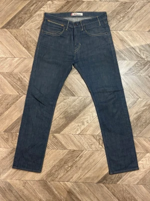 Levi's 504 Straight jeans - Säljer ett par klassiska mörkblå jeans från Levi's med rak passform och fem fickor. Jeansen har kontrastsömmar och en liten röd Levi's-tag på bakfickan. Tillverkade i slitstark denim, perfekta för en clean och tidlös look. Längd 105cm bredd 41cm