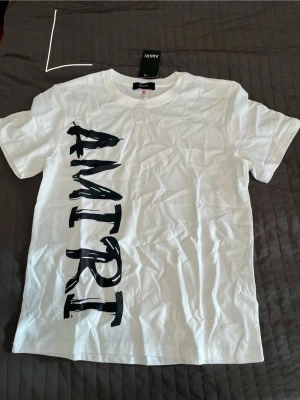 Vit AMIRI t-shirt med tryck - Snygg vit t-shirt från AMIRI med stort svart tryck längs ena sidan framtill. Klassisk rund hals och korta ärmar. Perfekt för dig som gillar streetwear och vill sticka ut med en stilren design. Gjord i mjuk bomull för skön känsla hela dagen.