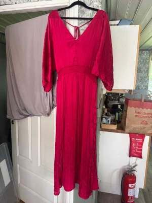 Röd långklänning med v-ringning - Lång klänning i krinklad satin med V-ringning fram och bak. Modellen har knytband i nacken, smock i midjan och slitsar i sidorna för en bekväm och smickrande passform. 3/4-ärmar. Tillverkad i 100 % polyester. Storlek 42/44
