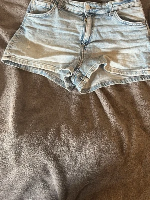 Ljusblå denimshorts med fickor - Säljer ett par ljusblå denimshorts med klassisk femficksdesign och dragkedja framtill. Shortsen har normal passform och är perfekta för varma dagar. Snyggt slitna detaljer och mjukt bomullsmaterial för en avslappnad look.