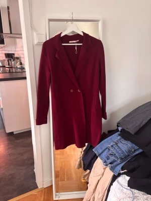 Vinröd lång kappa med knappar - Stilren vinröd kappa med dubbelknäppning och klassisk krage. Lång modell med raka ärmar och två fickor framtill. Perfekt för höst och vinter, ger en elegant look till din outfit. Materialet ser ut att vara mjukt och något tjockare, vilket passar bra till kallare dagar.