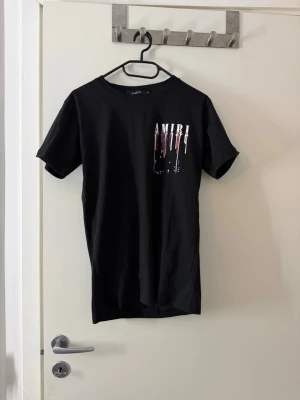 Svart AMIRI t-shirt med tryck - Säljer en svart t-shirt från AMIRI med coolt tryck fram och bak i vitt, rött och blått. T-shirten har korta ärmar och normal passform. Materialet känns mjukt och skönt, troligen bomull. Perfekt för dig som gillar streetwear och snygga detaljer.