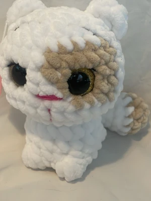 Vit och beige virkad kattmjukis - Supergulligt handvirkat gosedjur i form av en katt med stora gula ögon med glitterdetaljer. Katten är vit med beige fläckar och har rosa broderad nos och mun. Tillverkad i mjukt, fluffigt garn som känns mysigt att krama. 17cm