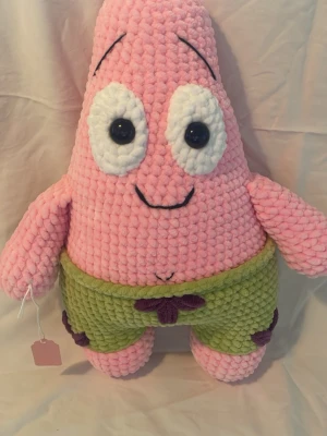 Stickad Patrick Star mjukisdjur - Supermjuk och gosig stickad Patrick Star-figur från Svampbob Fyrkant. Den är rosa med stora vita ögon, svarta detaljer och gröna shorts med lila blommor. Perfekt som prydnad eller kramkompis för alla som gillar tecknade serier. 33 cm
